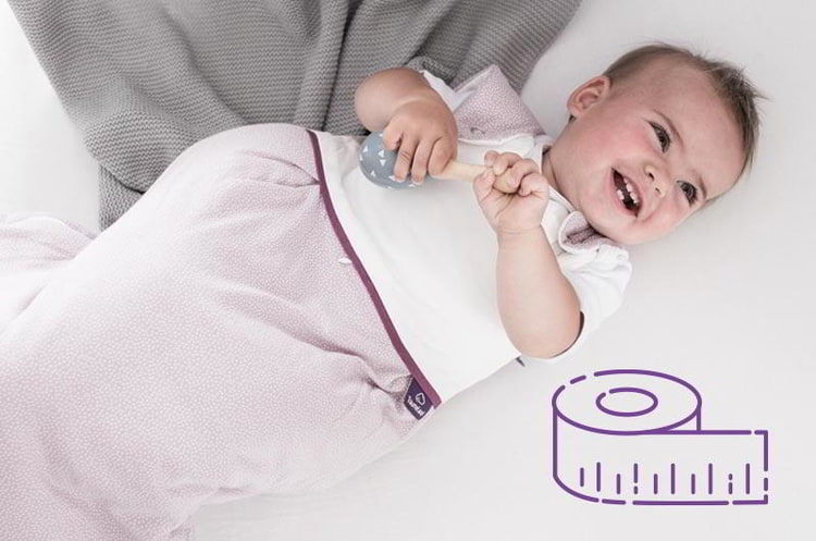 Bebé en saco de dormir rosa Träumeland en la cama con ilustración de una cinta métrica Bebé en saco de dormir rosa Träumeland en la cama con ilustración de una cinta métrica