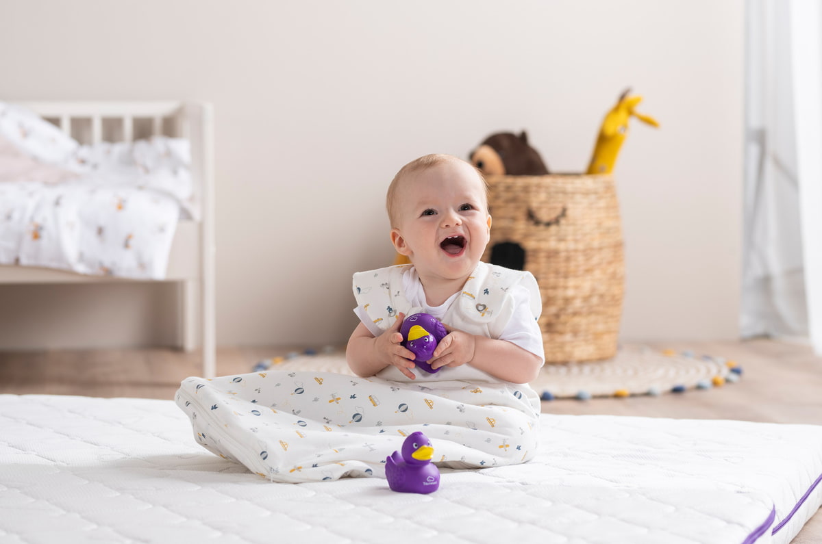Bebé en saco de dormir Träumeland sentado en colchón con pato chirriante Bebé en saco de dormir Träumeland sentado en colchón con pato chirriante